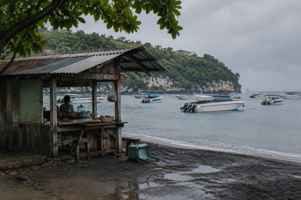 A wooden kiosk selling fast boat tickets beside a Bali harbour with boats at anchor