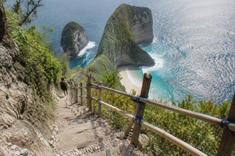Narrow cliff trail with wooden steps and a bamboo handrail descending toward a small white sand beach between tall rock walls