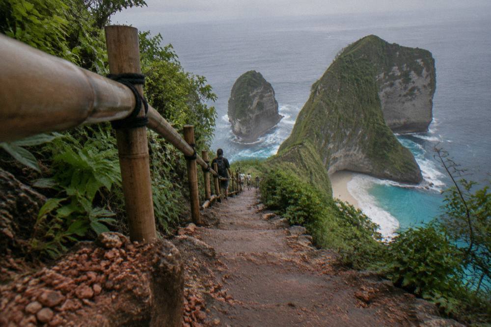 Steep dirt trail with wooden and bamboo railings descending the cliff side above Kelingking Beach on Nusa Penida