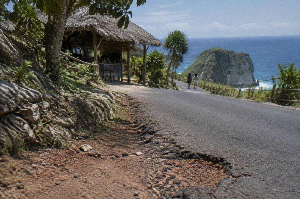 Steep rocky trail descending to Kelingking Beach with a low bamboo handrail and ocean beyond