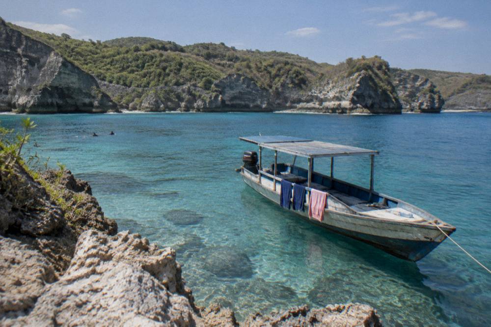 Snorkel boat anchored over clear shallow water near a rocky Nusa Penida shoreline