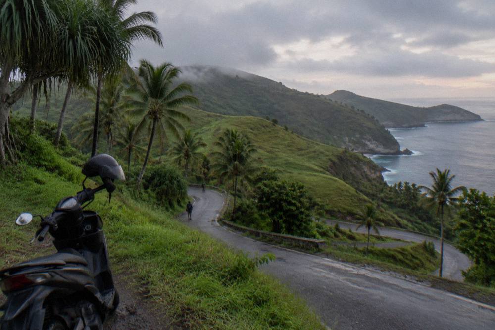 A steep narrow asphalt road winding down a green hillside toward the coast