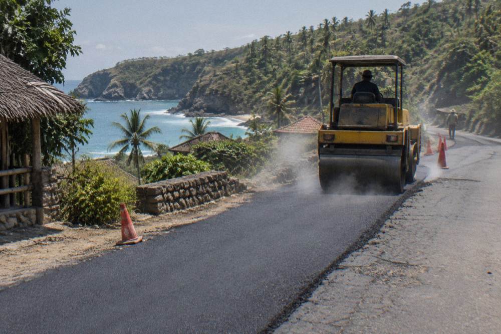 Roadworks machinery and fresh asphalt on a coastal road beside green hills