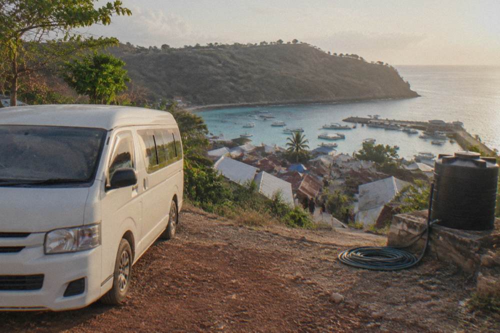 An air-conditioned minivan parked on a coastal road above a turquoise bay